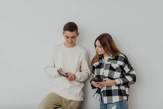 Teenage boy and girl using smartphones while leaning against a white wall indoors. How COPPA and California's design code change social apps for US teens
