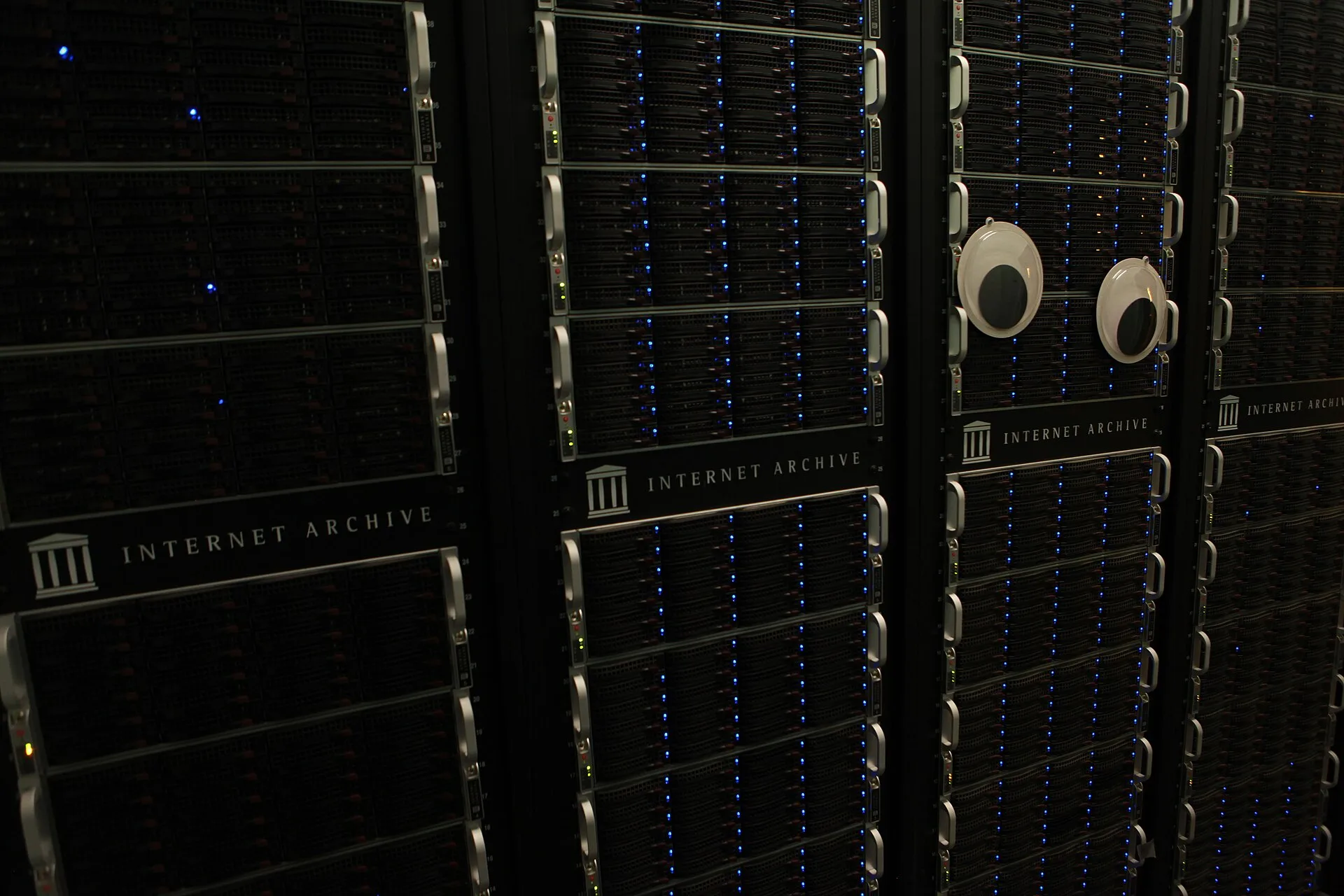 An isle of server racks in an Internet Archive data center, with large googly eyes on the front of one rack. The ethics and mechanics of archiving internet culture properly