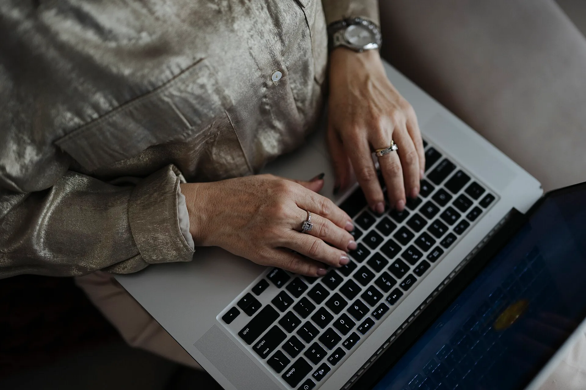 A woman sits on a sofa using a laptop. Her hands are on the keyboard. The room is light and the time looks like afternoon. She wears rings on her fingers. Social media slang statistics 2027: how to source them